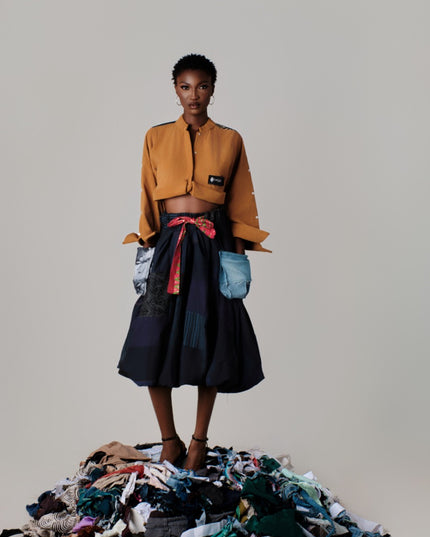 Person wearing a mustard jacket and dark skirt standing on a pile of clothes against a plain background