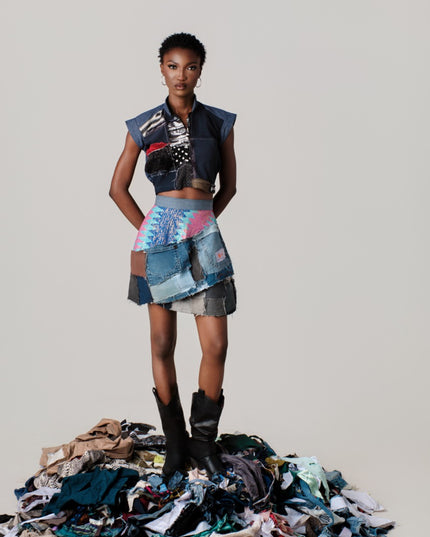 Model wearing a colorful, patchwork outfit standing on a pile of fabric scraps against a plain background.
