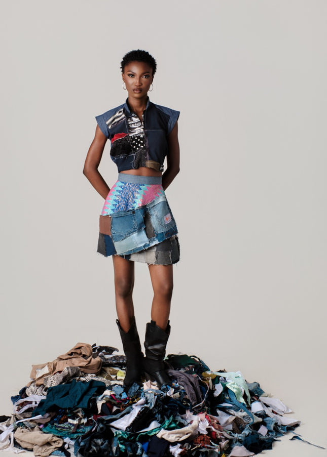 Model wearing a colorful, patchwork outfit standing on a pile of fabric scraps against a plain background.